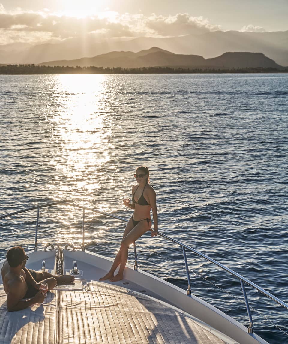 A couple in swimwear relaxing on the bow of a boat, wine glasses in hand and the setting sun glinting off the endless ocean.