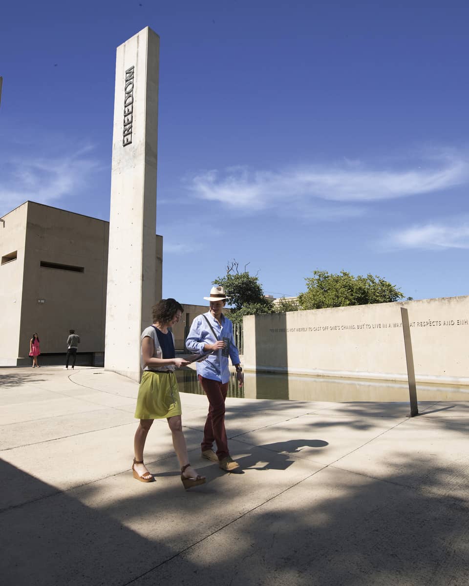 Man and woman walk on path past white pillars, wall with Nelson Mandela quote