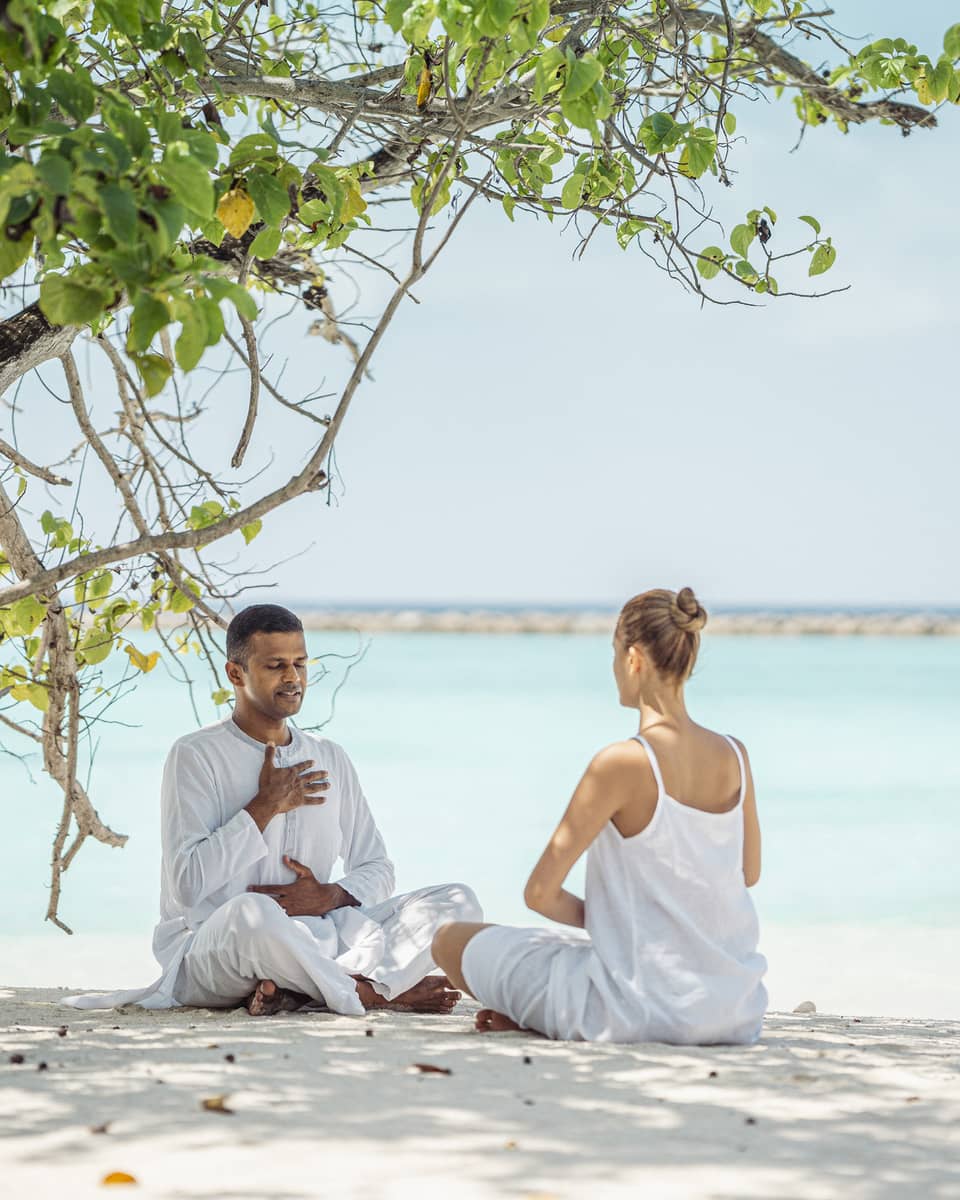 Dr. Arun K. Tomson practises breathing exercises with female guest on the beach under the shade of a tree