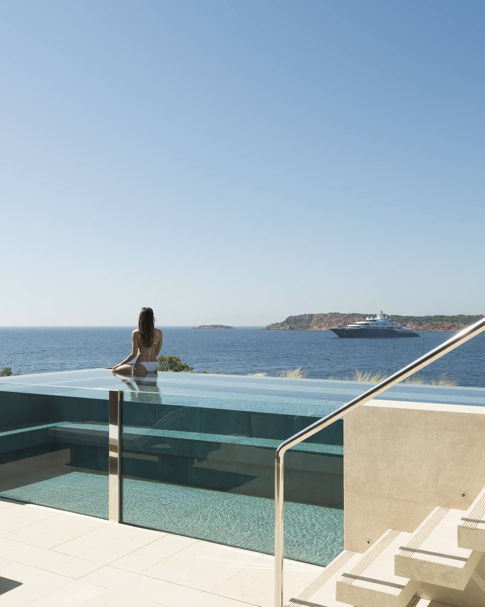 Terrace with staircase and glass infinity pool, a woman wearing white bikini in pool looks out to sea