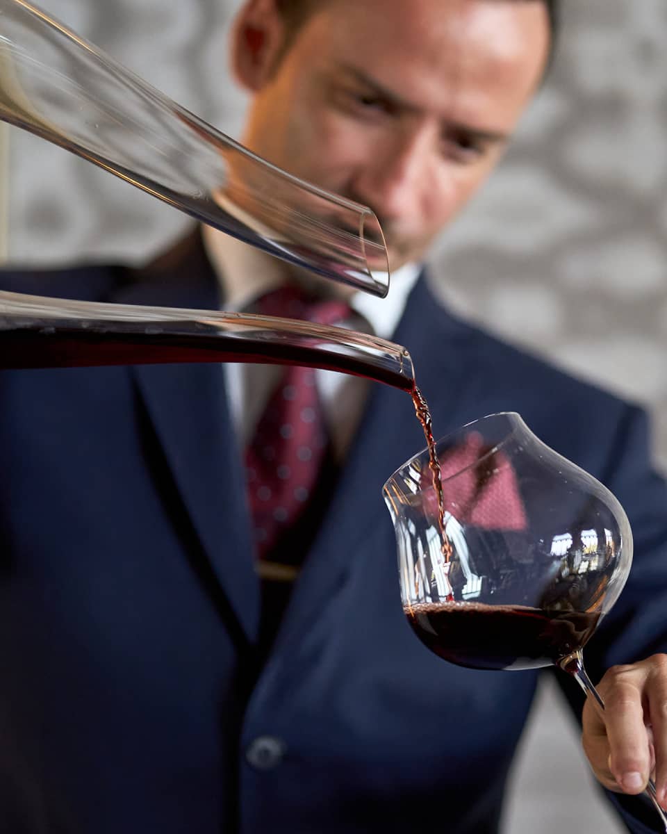 A person in formal attire carefully pouring red wine from a decanter into a glass.