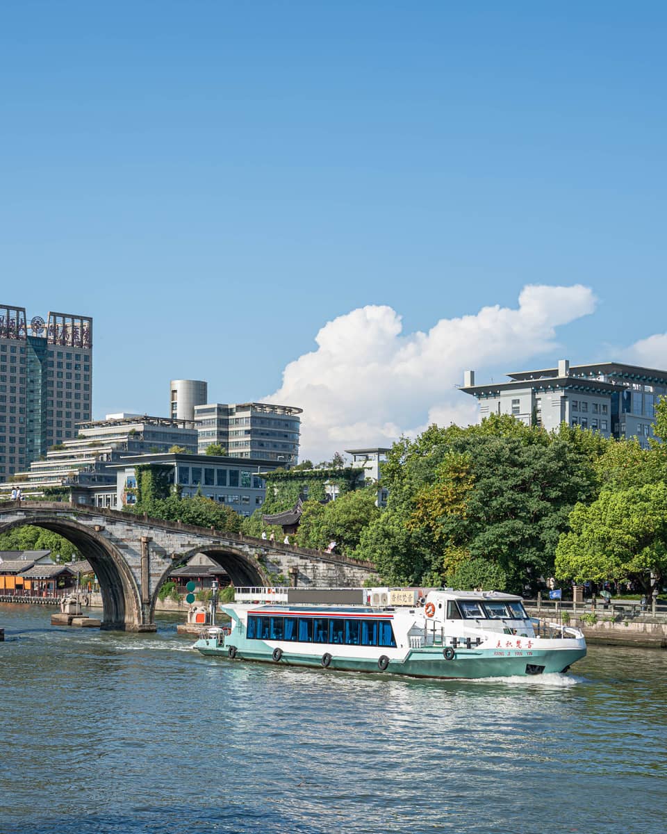 A tour boat passes under the centre arch of a historic stone bridge spanning a canal flanked by trees and modern buildings.