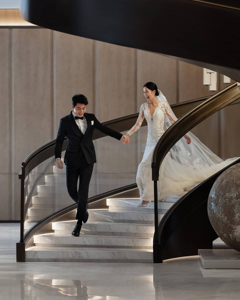 A smiling bride and groom walk hand in hand down the stairs at Four Seasons Hotel Dalian.