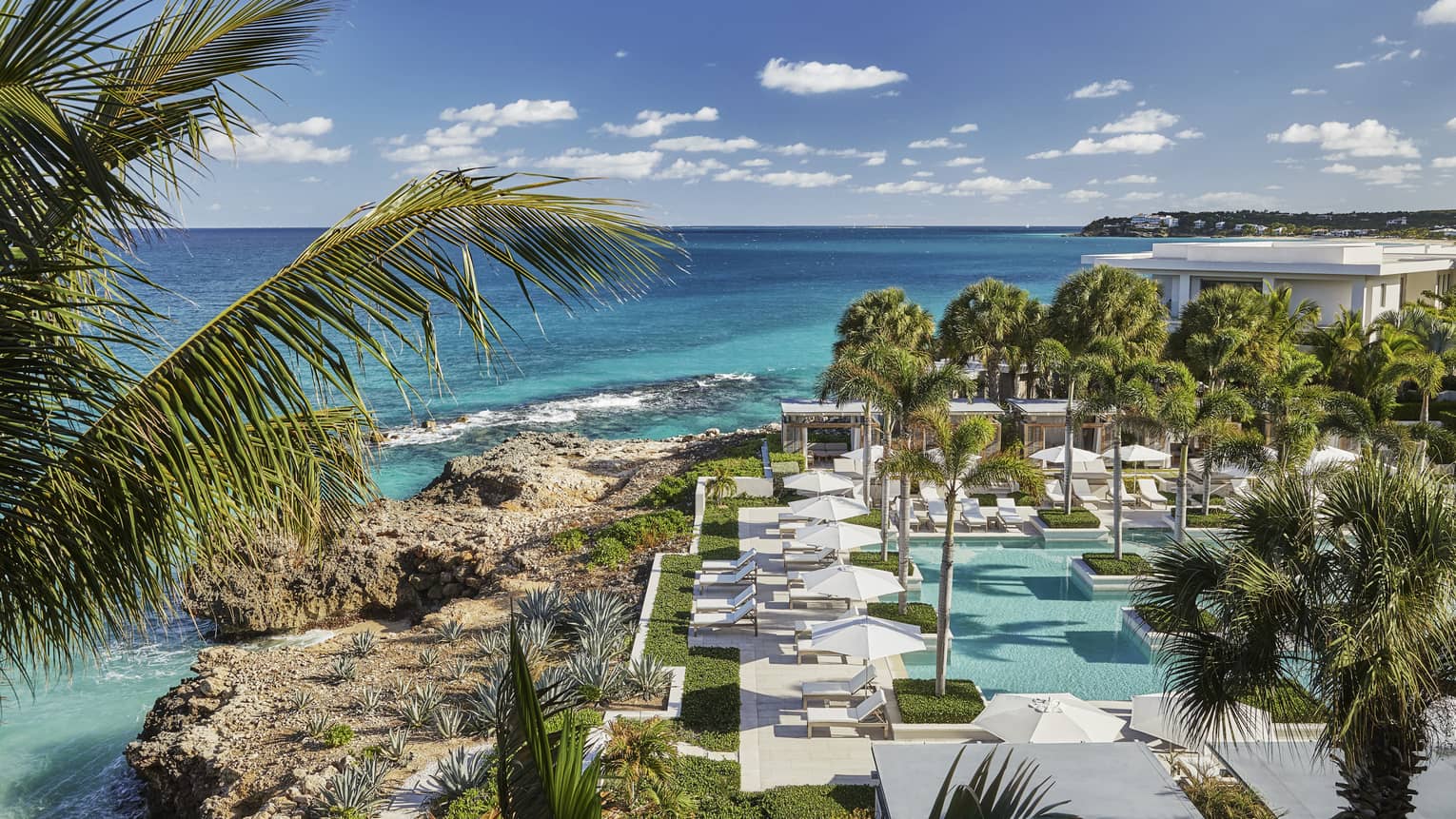 Looking down over Anguilla outdoor pool, patio, large rocks along blue-green ocean waterfront, blue sky