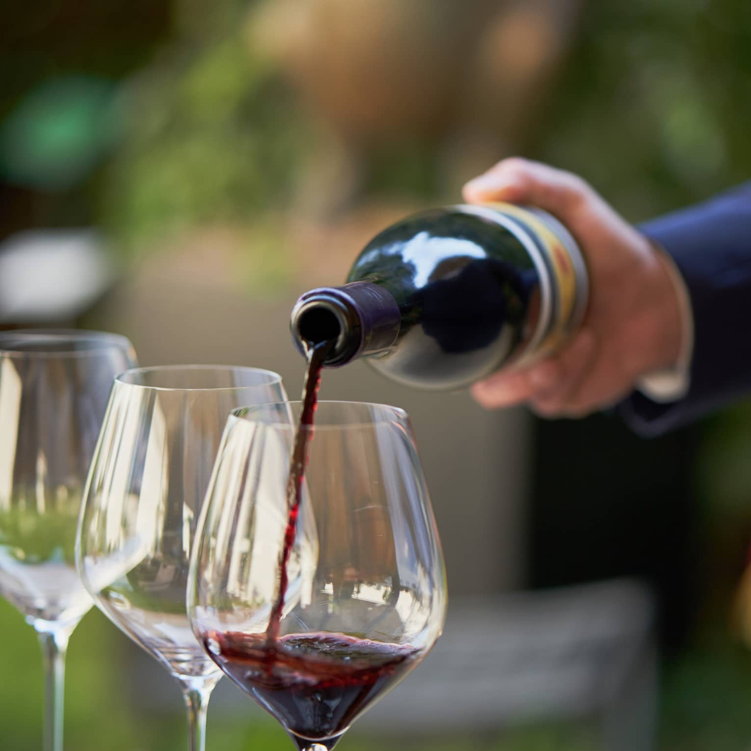 A close up of hands pouring a bottle of red wine into a clear wine glass that rests on a wooden tray, outdoors