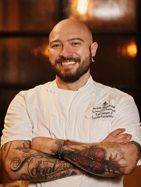 A smiling chef wearing a white Four Seasons uniform stands with arms crossed.